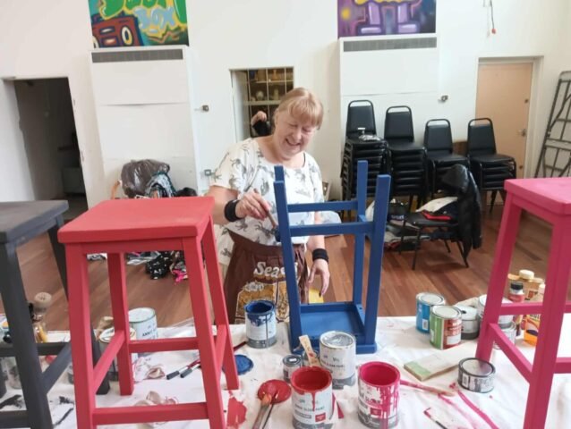 women painting a school stool blue with two other stools next to her that have been painted red